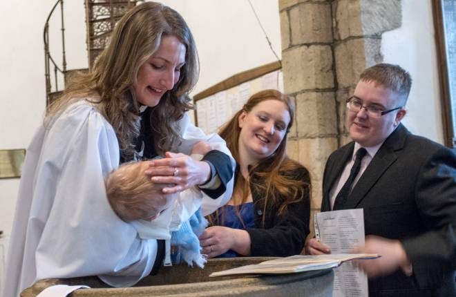 female vicar baptising baby in font, parents standing beside it smiling 
